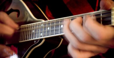 Primer plano de una mano pisando un acorde en el m&aacute;stil de una mandolina: se ven las ocho cuerdas agrupadas en cuatro &oacute;rdenes dobles, los trastes y los dedos presionando dos cuerdas a la vez