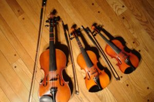 Tres violines de estudio de tamaños 4/4, 1/8 y 1/10 con sus arcos sobre un suelo de madera