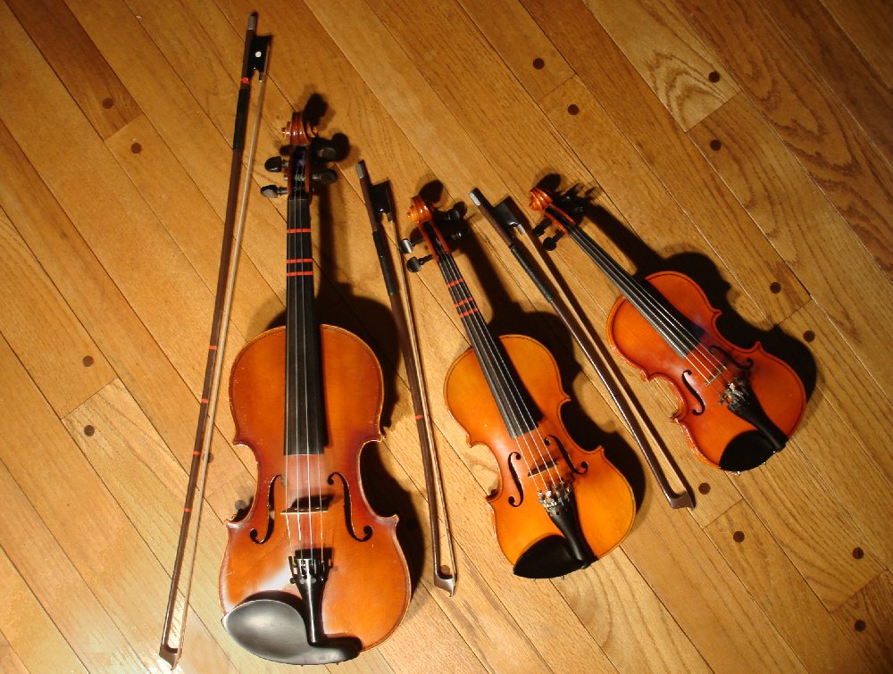 Tres violines de estudio de tamaños 4/4, 1/8 y 1/10 con sus arcos sobre un suelo de madera